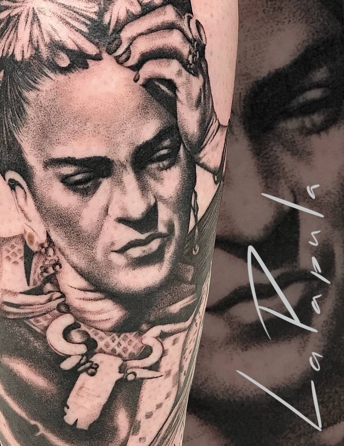 Black and grey realistic tattoo of a woman with flowers in her hair and a hand resting on her forehead, with a blurred detail of another face behind it.