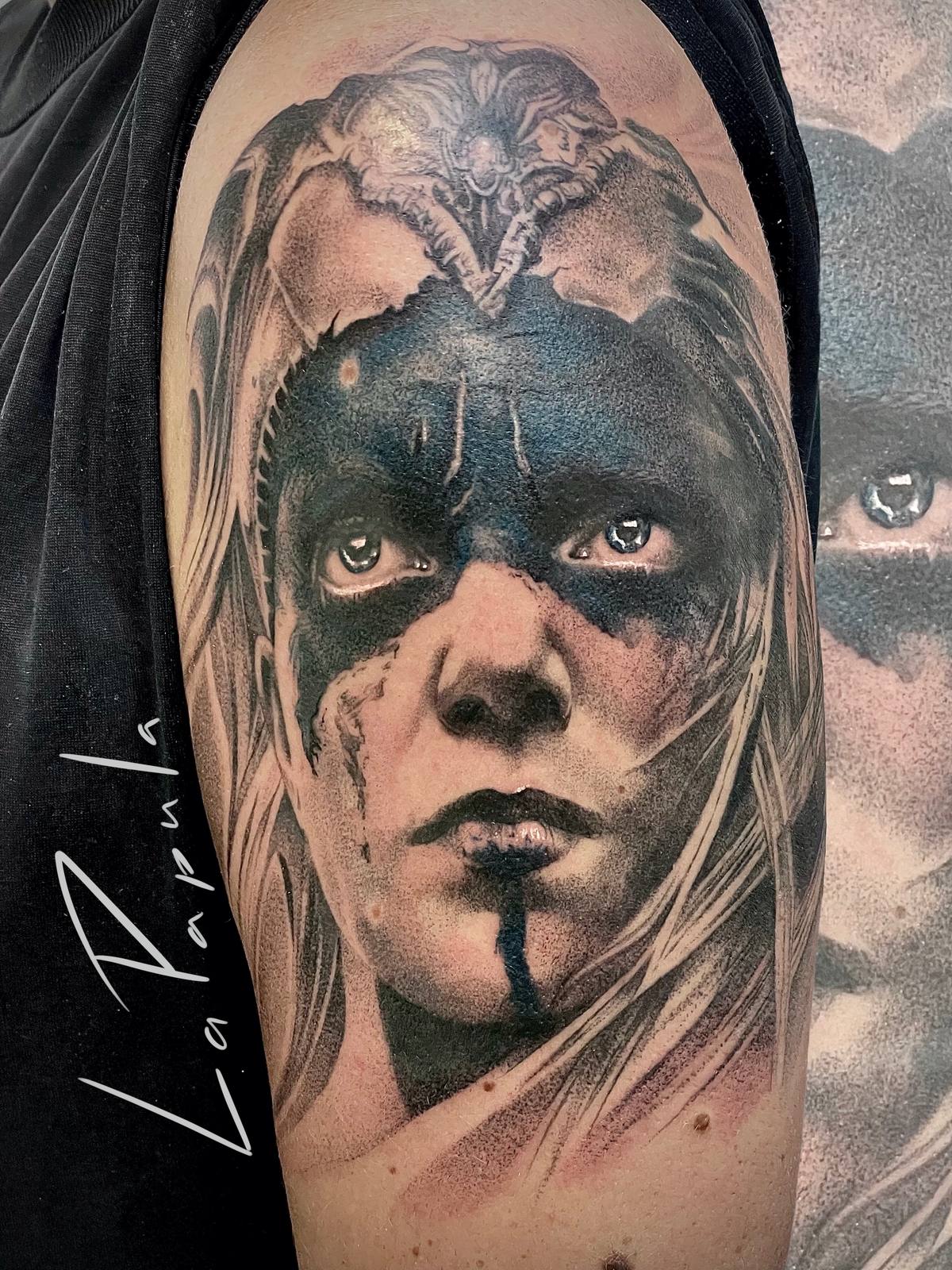 Realistic tattoo of a woman's face with dark paint around the eyes and long light hair, on the upper arm.